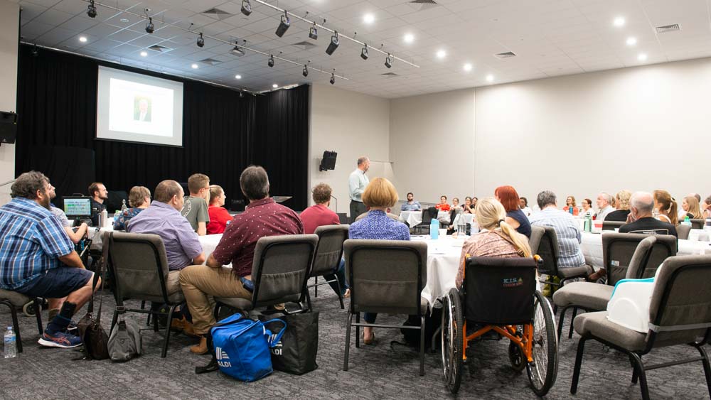 A diverse group of people, some in wheelchairs, sitting around tables and watching a man present