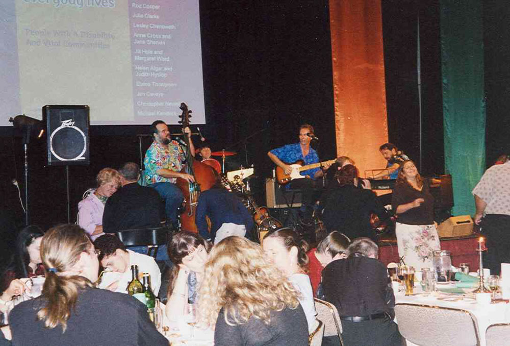 A band is playing music on stage to a crowd of people sitting at tables full of beer and food.  Projected behind them is the poster from the conference.