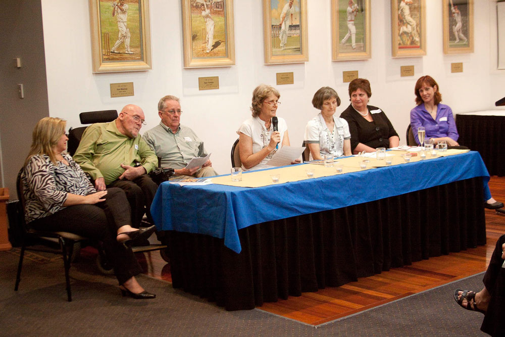 A group of 7 people sitting at a table with paintings of cricket players behind them.  One of the people is in a wheelchair and another is speaking in to a microphone.