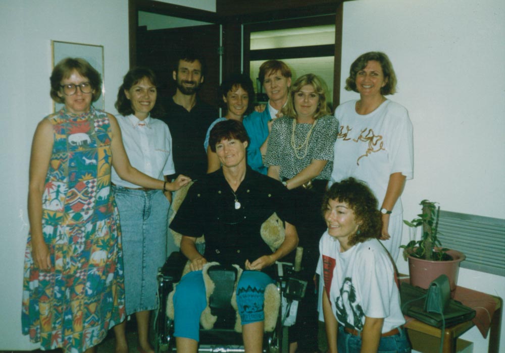 A group of women sitting and standing in an office, including a woman in a wheelchair with a main the background