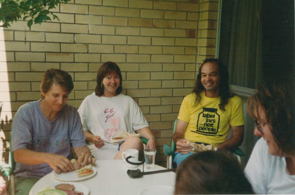A group of people sitting around a table outside smiling. One man is wearing a shirt that says 'label jars, not people'