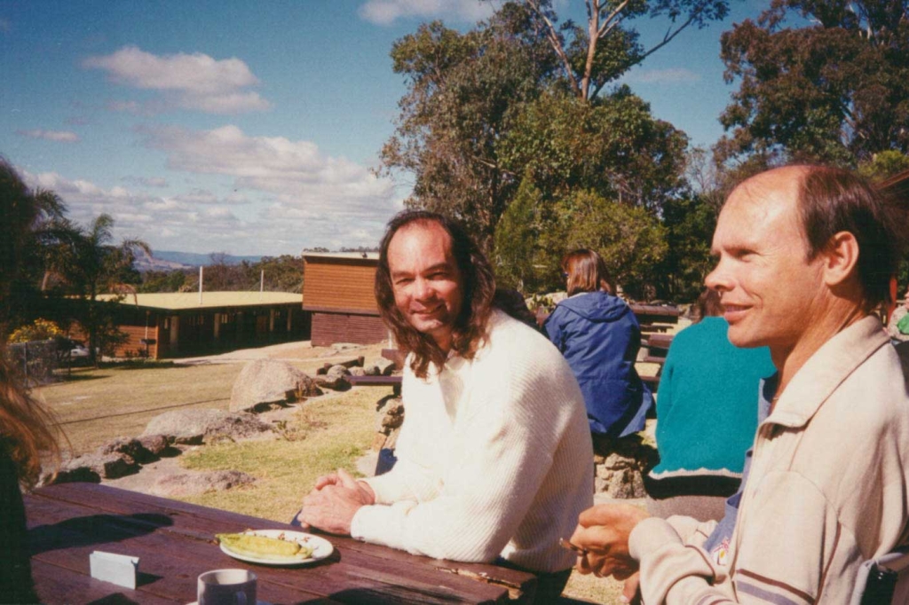 2 men sitting outside in the sun, looking at a nice view under trees