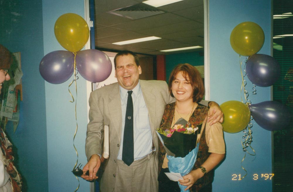 A man and a woman laughing and hugging, surrounded by balloons and streamers in a party scene