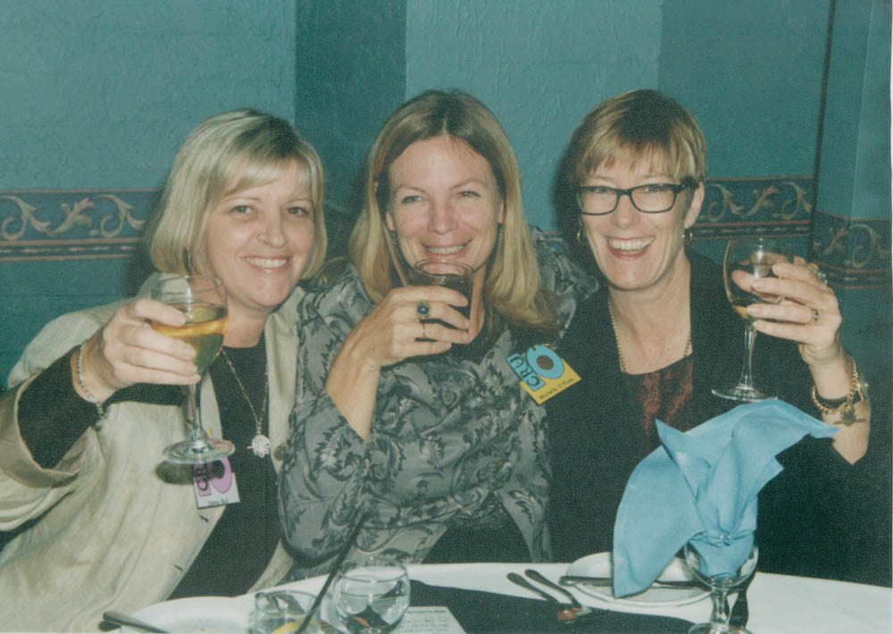3 women sitting together, smiling, toasting the camera with glasses of wine.