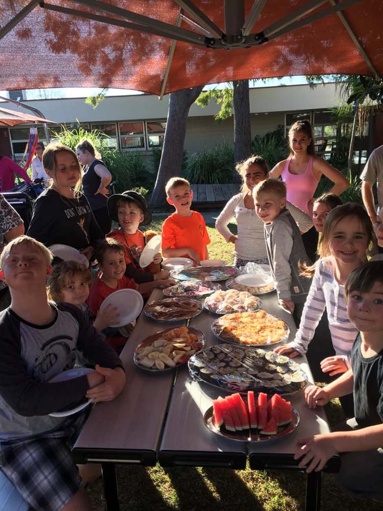 A group of kids sitting around a picnic table filled with food they have prepared.  Many of the children have disability.