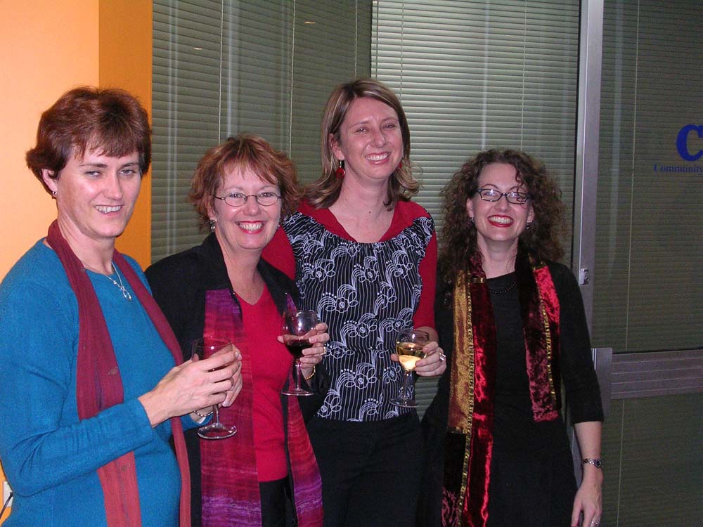 Four women standing together with glasses of wine smiling in an office, with the letters C R U on a glass wall behind them