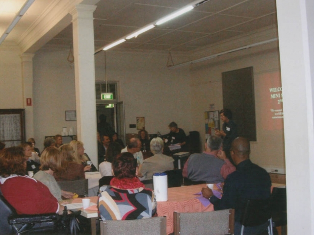 A group of people, some in wheelchairs, sitting at tables looking at a presenter.  Behind the presenter a slide is projected and it read 'welcome to CRU's mini summit 2009'.