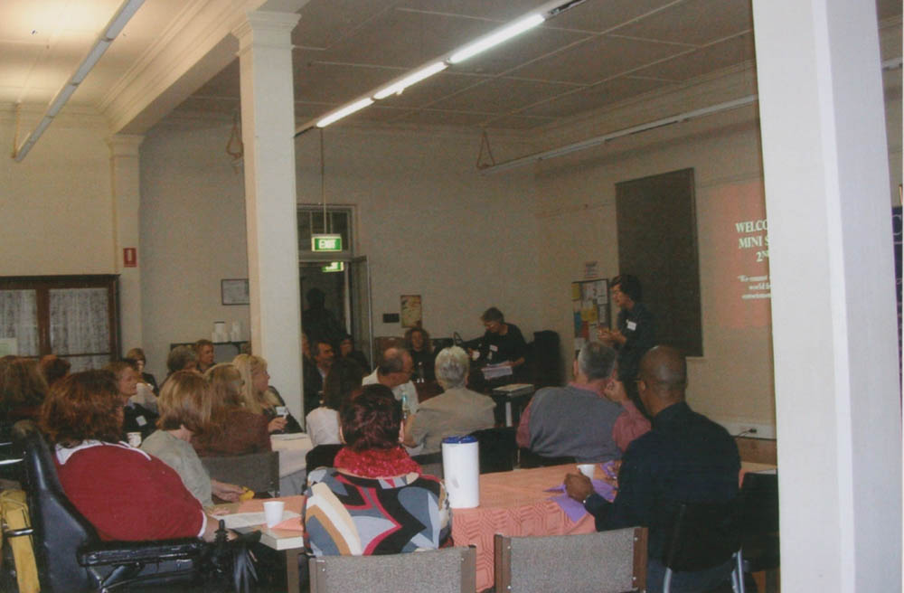 A group of people, some in wheelchairs, sitting at tables looking at a presenter. Behind the presenter a slide is projected and it read 'welcome to CRU's mini summit 2009'.
