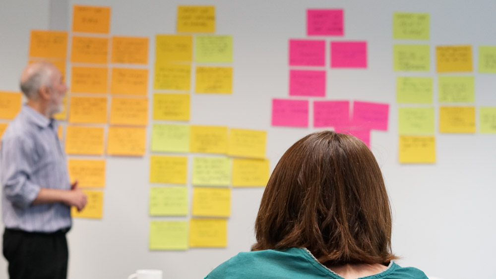 In the background a man looks at notes on a wall, while in the foreground is the back of a woman's head.