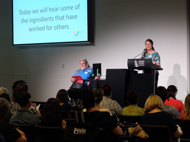 A woman stands on stage and speaks at a lectern and introduces the speaker who is beside her.  He is in a wheelchair and wears a Q D N shirt.