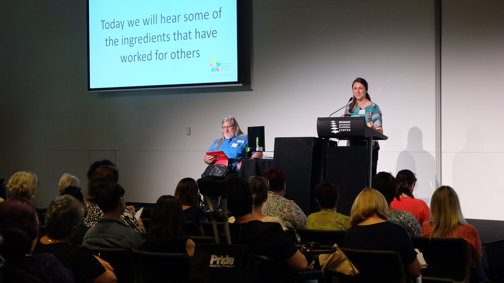 A woman stands on stage and speaks at a lectern and introduces the speaker who is beside her. He is in a wheelchair and wears a Q D N shirt.