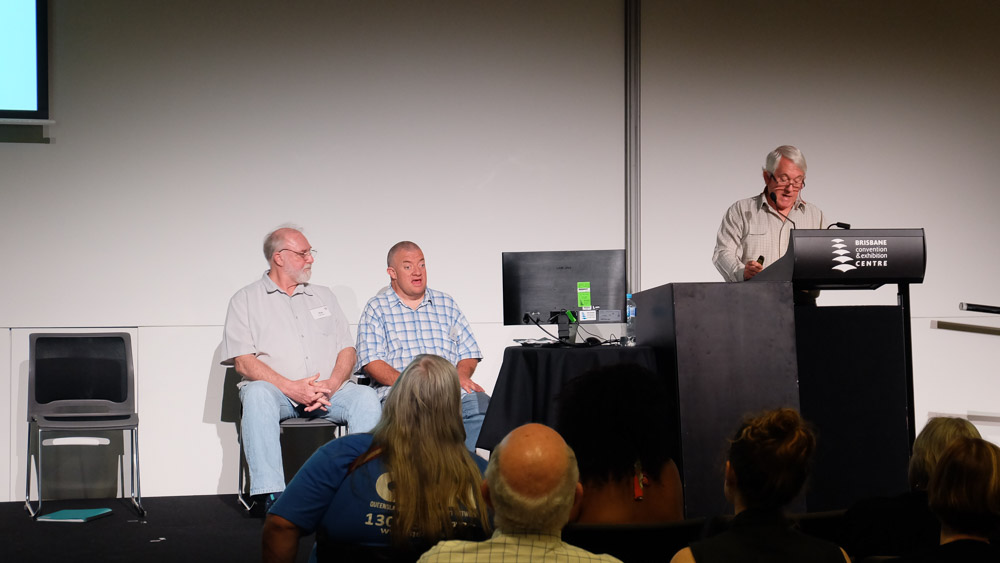 3 men on stage at the brisbane convention and exhibition centre.  One is a son and his support worker, watching the father speak at the podium.