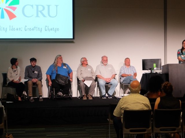 A cru staff member speaks in to a microphone at a lectern. She is on stage with a group of presenters - people with disability and their friends/family who are supporting them