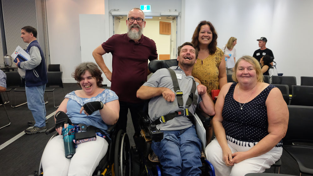 3 women and 2 men laughing and smiling at a cru event, surrounded by empty chairs at the end of the day. Two of the people are in wheelchairs.