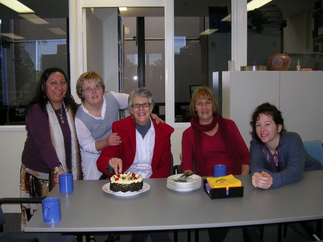 5 women sitting and standing together in an office.  One of the women is cutting a birthday cake