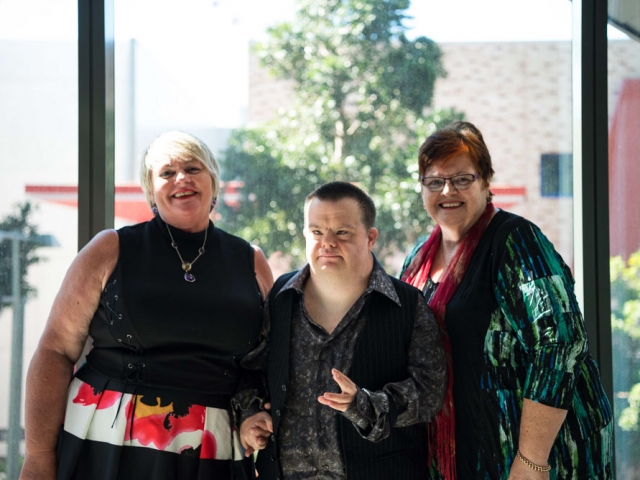 3 people standing together in front of a large window.  one of them has a disability.