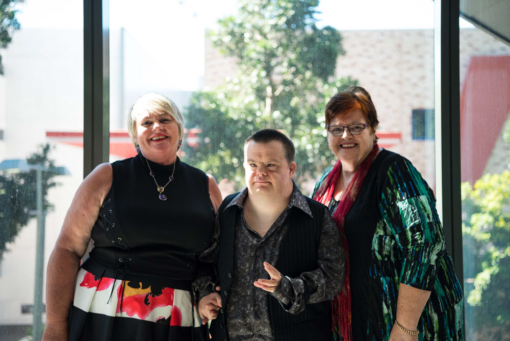 3 people standing together in front of a large window. one of them has a disability.