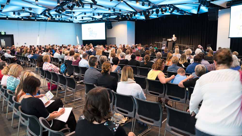 A large group of people in an auditorium, taking notes as they look to the presenter