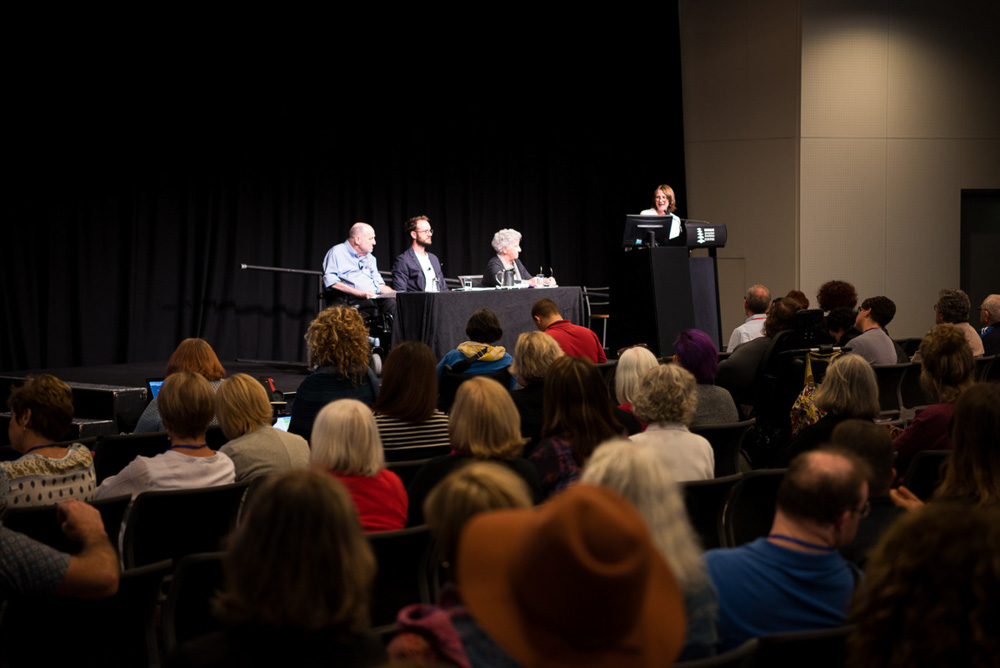 4 people on stage at the front of a large group at a conference. one woman at a lectern looks at three panel members. one of the panel has a disability