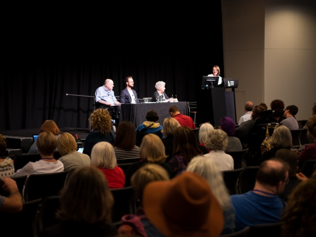 4 people on stage at the front of a large group at a conference.  one woman at a lectern looks at three panel members.  one of the panel has a disability