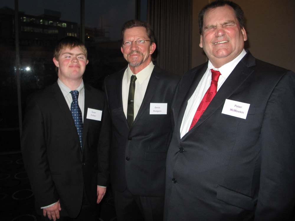 3 men in suits standing and smiling, all wearing name badges. Two of them have a disability