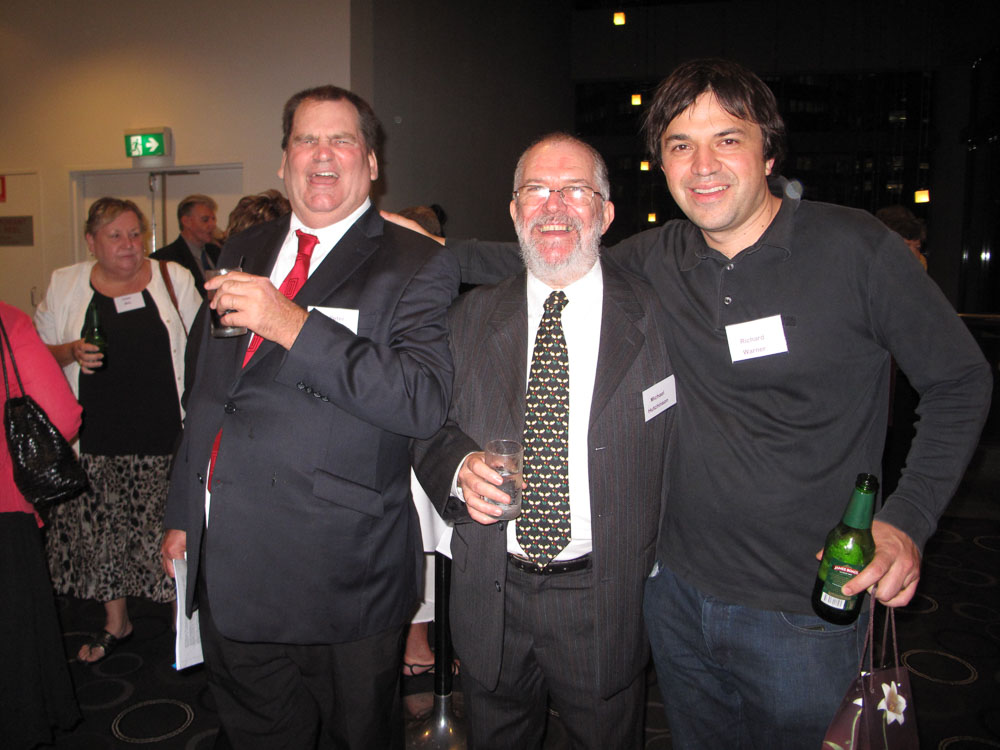 3 men standing together smiling at a conference. They are dressed in suits, drinking and enjoying themselves.
