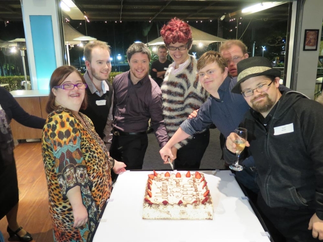 7 young adults with disability, all well dressed and smiling at a celebration at a fancy restaurant.  They all have their hands on a knife cutting a cake that has the words 'celebrating 35 years of QPPD'