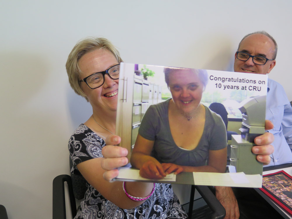 A woman with disability holds a book that has a photo of her on its cover. It has the words 'congratulations on 10 years at cru' on its cover. Behind her a colleague looks at her smiling warmly.