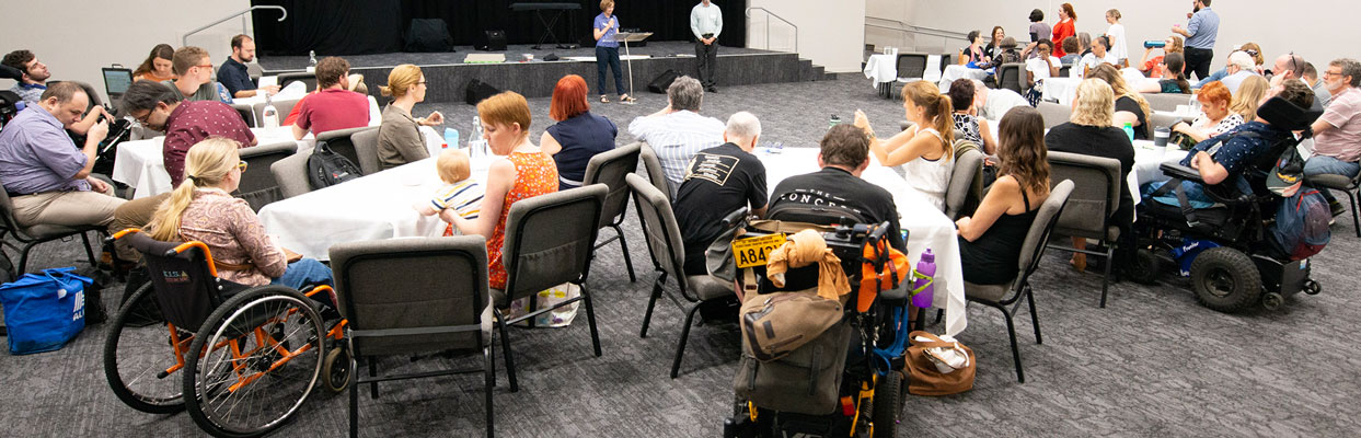 A photo of a cru leadership retreat. It's a diverse room of people with disability, friends, family and workers - doing activities around a series of tables.