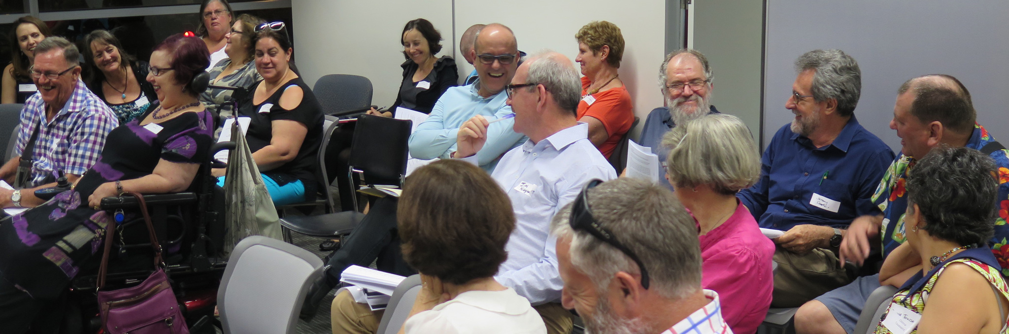 A group of people, some with disability, sitting together in a gathering laughing and smiling warmly together.