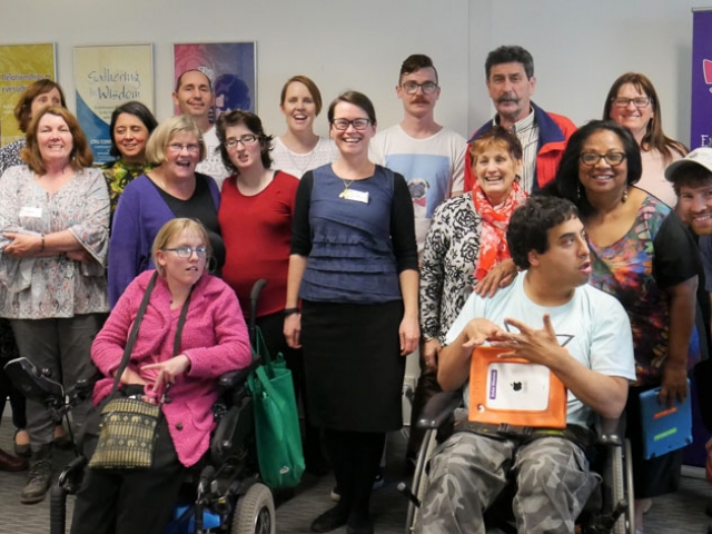 A diverse group of around 25 people - many of whom have a disability - are in the cru office and smiling at the camera.