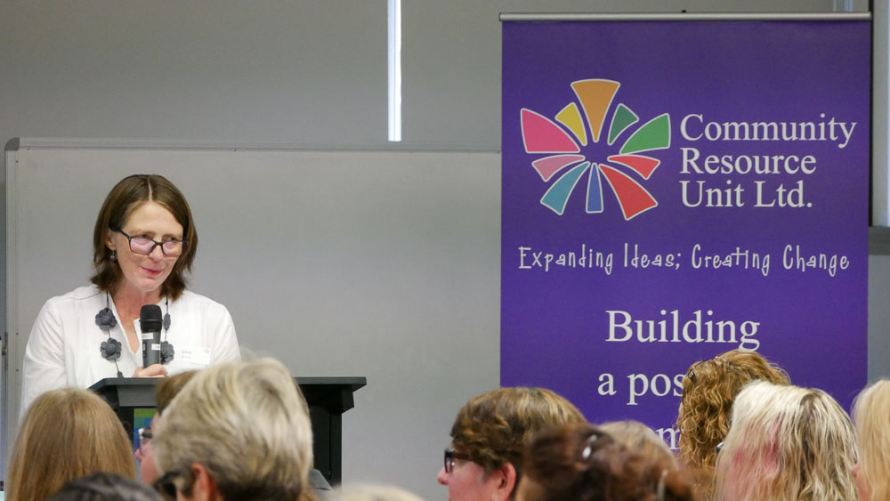 Lisa Bridle from cru presenting in front a group of people with the cru banner to her right.  The banner reads community resource unit ltd.  Expanding ideas; creating change