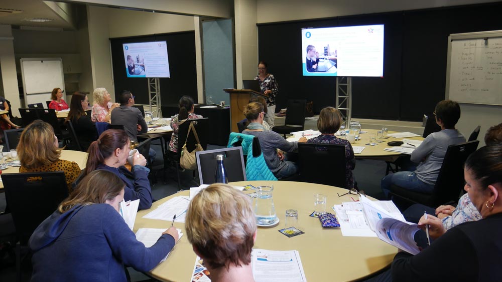 Lindie from cru presenting to a group of parents sitting around tables, writing notes and looking at her present