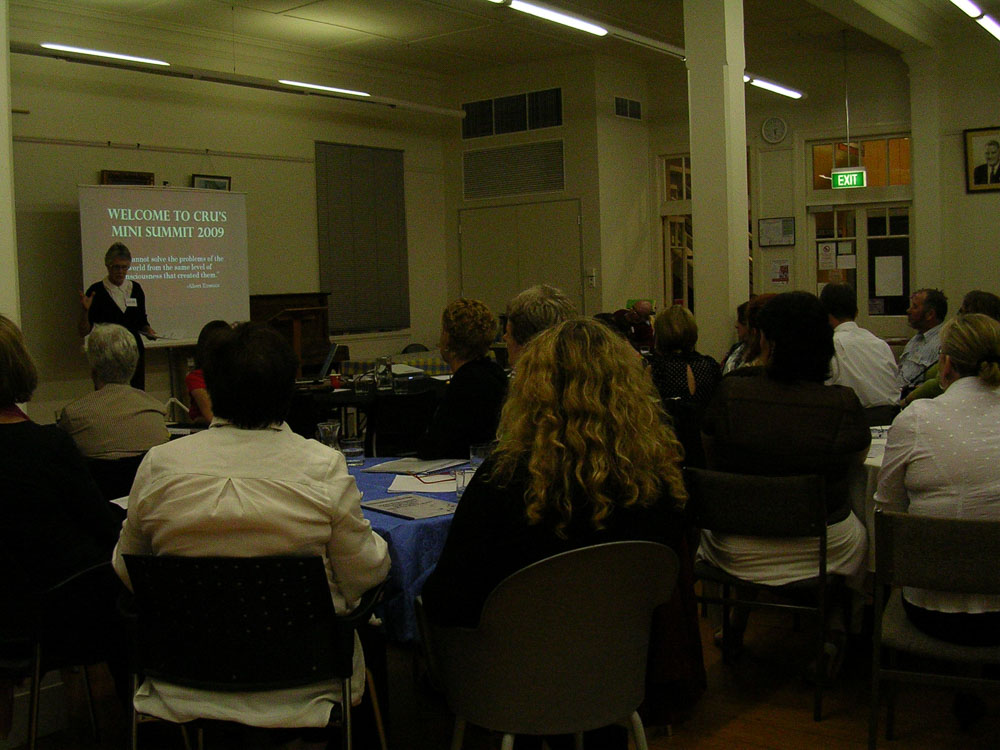 A group of people sitting at tables looking at a presenter. Behind the presenter a slide is projected and it read 'welcome to CRU's mini summit 2009'.