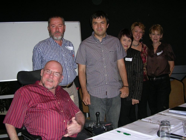 6 people standing together behind a table covered in folders and with a microphone on it.  One of the men is in a wheelchair