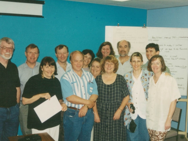 A dozen men and women standing in from of a whiteboard smiling and standing close together