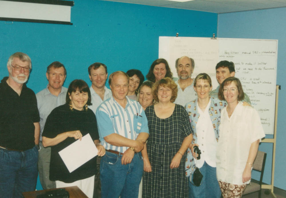 A dozen men and women standing in from of a whiteboard smiling and standing close together