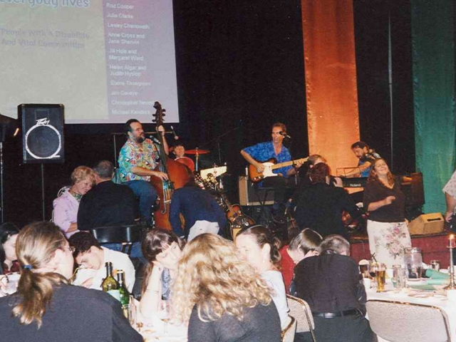 A band is playing music on stage to a crowd of people sitting at tables full of beer and food.  Projected behind them is the poster from the conference.