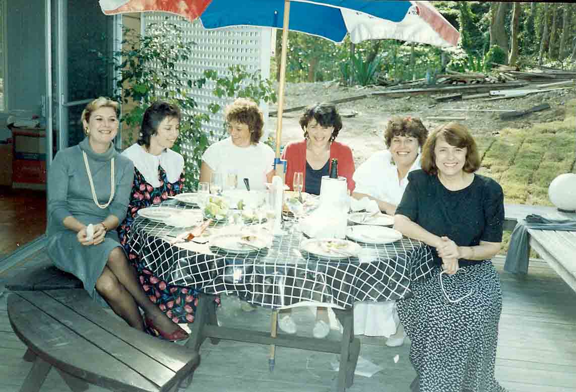 6 women sitting at a table outside, having eaten, drinking wine and enjoying themselves.