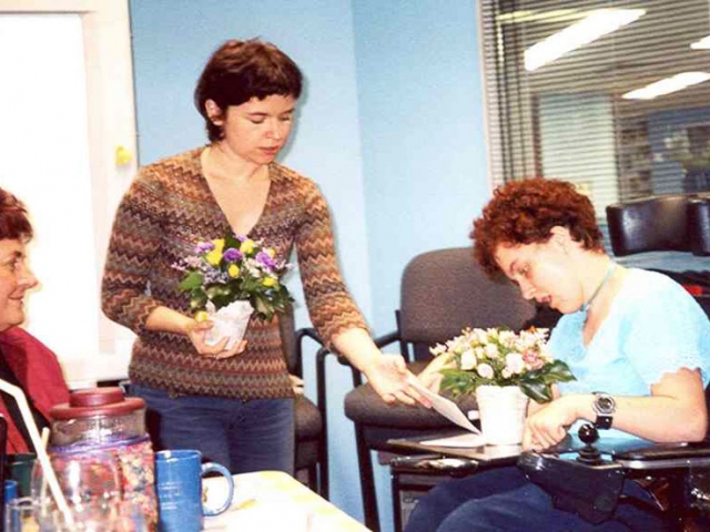 A woman in a wheelchair is assisted to give two bunches of flowers and a card to another woman sitting and facing her