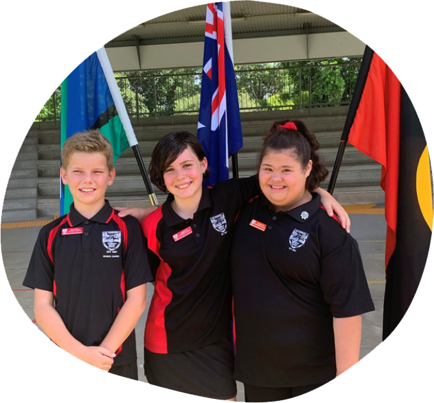 3 children in school uniforms standing in front of three flags - Australian, Torres Strait and our indigenous flag. One of the children has a disability