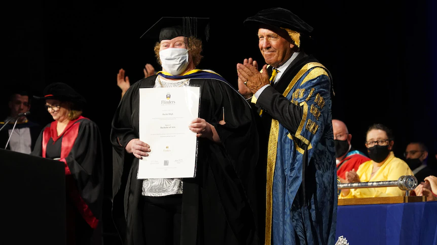 A man wearing graduation gown, cap and a mask holding graduation certificate with people around clapping.