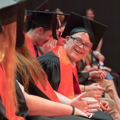 Young man smiling and wearing graduation ceremony gown and sitting alongside his peers.