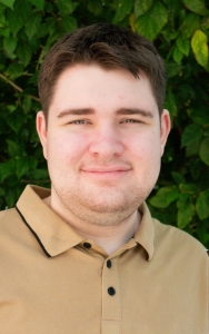 Cooper smiling, he has short brown hair and is wearing a collard shirt. Leaves in background.